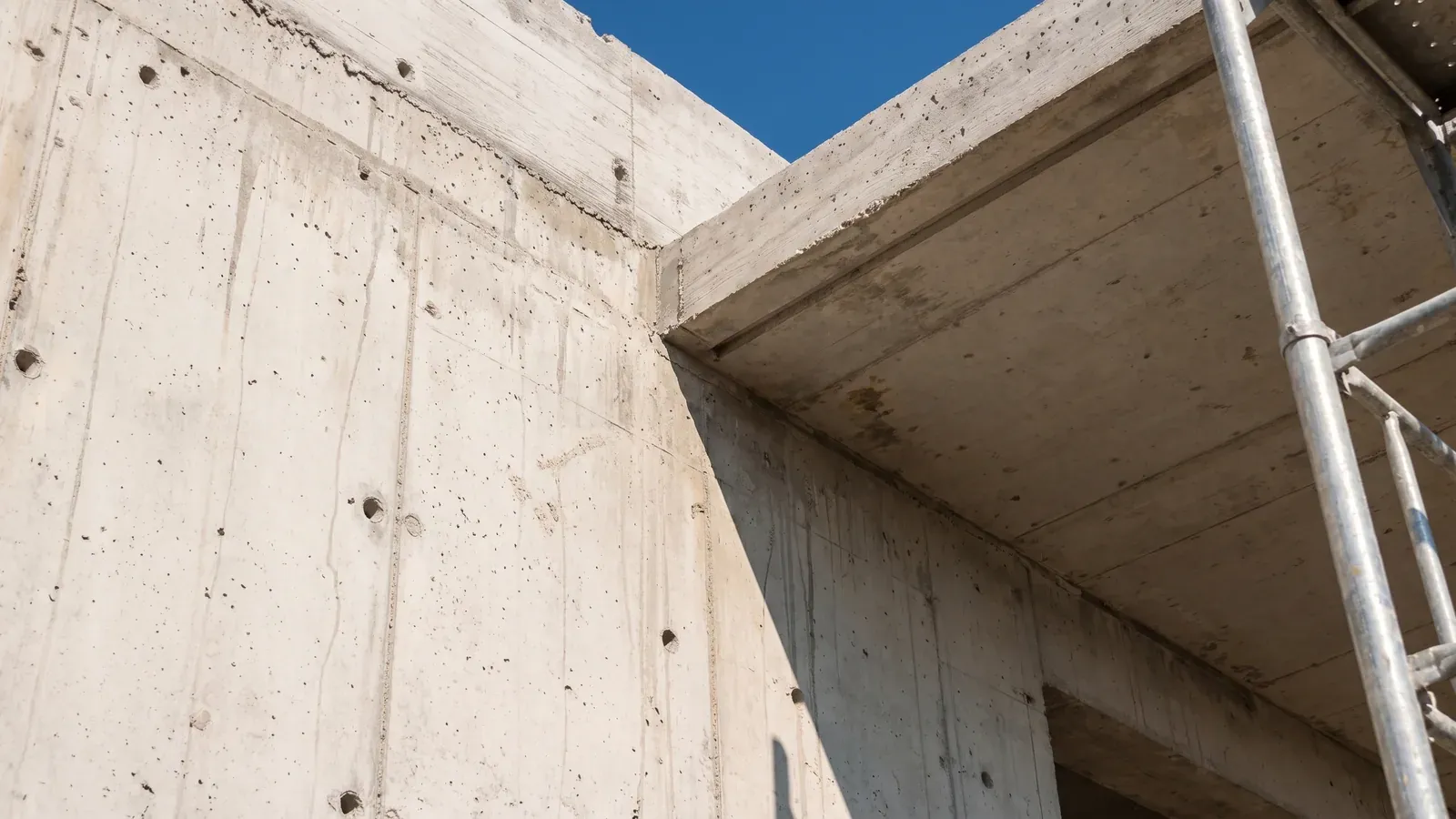 Villa şantiyesinde brüt beton duvar ile konsol teras döşemesinin birleştiği köşe; kalıp tahtası dokusu, düzenli bağ deliği ızgarası ve damlalık kanalı